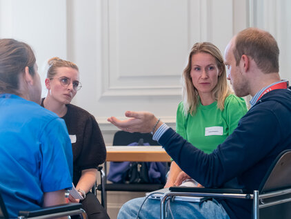 Vier Personen sitzen in einem Kreis und diskutieren. Eine Person spricht gestikulierend, während die anderen aufmerksam zuhören. Alle Teilnehmer tragen Namensschilder und zeigen ein interessiertes Gesicht. Die Atmosphäre wirkt offen und kooperativ.