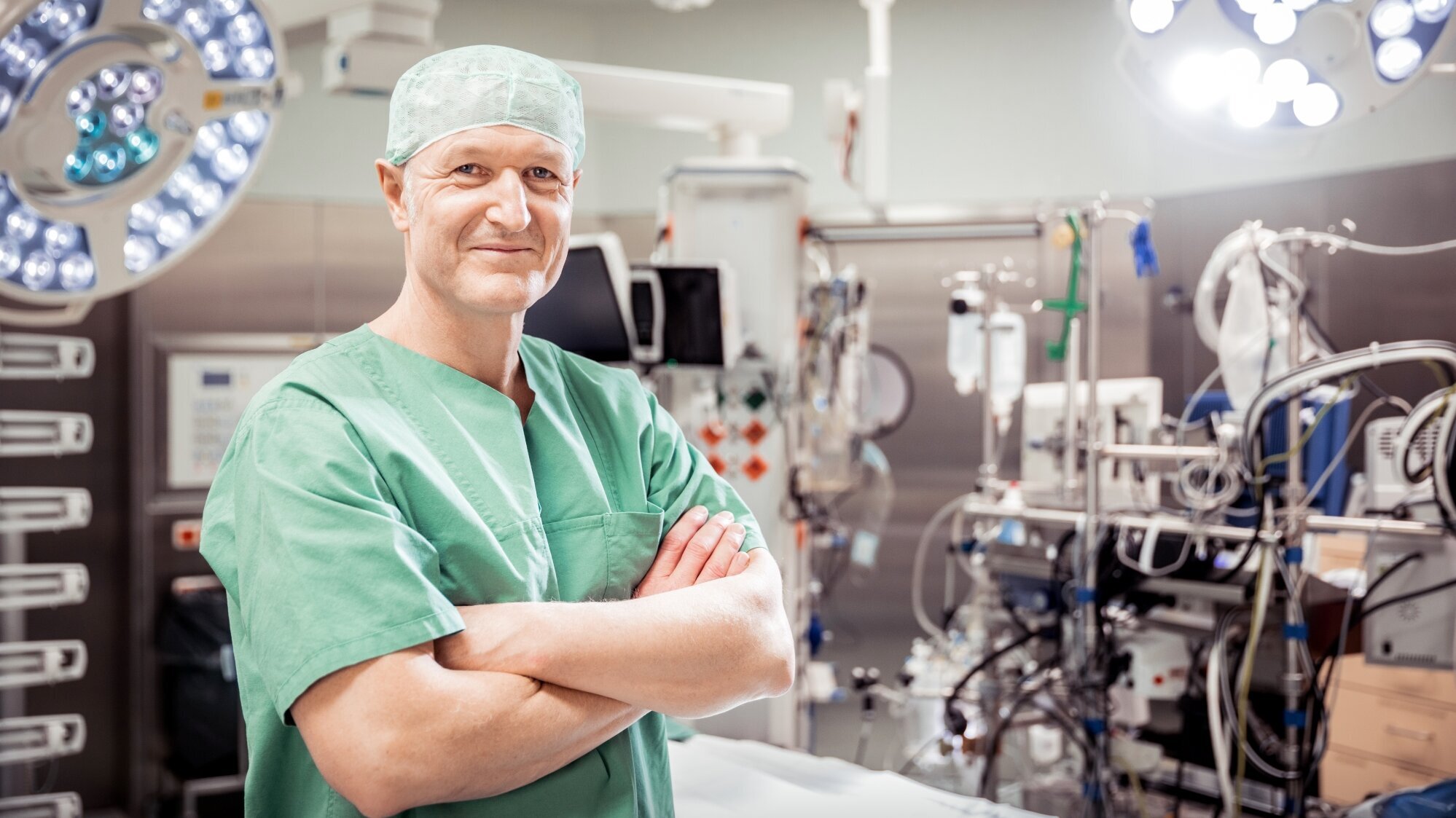 Ein lächelnder Chirurg in grüner OP-Kleidung steht mit verschränkten Armen in einem Operationssaal. Im Hintergrund sind medizinische Geräte und chirurgische Lichter zu sehen. Der Raum ist bereit für einen Eingriff.