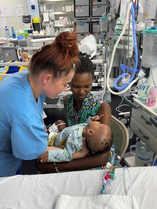 Eine Pflegekraft beugt sich zu einer Mutter, die ein kleines Baby in den Armen hält. Beide Frauen schauen freundlich, während das Baby die Hand der Mutter greift. Im Hintergrund sind medizinische Geräte zu sehen, die auf eine intensive Pflegeumgebung hinweisen.