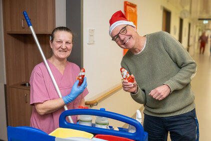 Eine lächelnde Pflegekraft in rosa Uniform hält einen Schokoladen-Weihnachtsmann. Neben ihr steht ein Mann mit einer roten Weihnachtsmütze, der ebenfalls einen Schokoladen-Weihnachtsmann in der Hand hält. Im Hintergrund ist ein Krankenhausflur sichtbar.