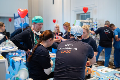 Eine Gruppe von Fachkräften in medizinischer Kleidung arbeitet gemeinsam an einem Tisch. Sie diskutieren und lernen in einem Ausbildungskontext. Auf ihren T-Shirts steht "Team Ausbildung DHZC". Im Hintergrund sind einige Personen und Dekorationen zu sehen, die das freundliche Ambiente unterstreichen.