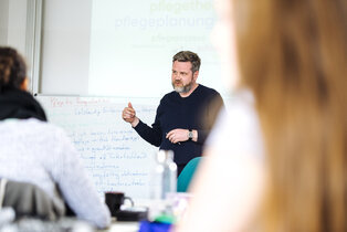 Ein Dozent steht vor einer Tafel und erklärt Themen zur Pflegeplanung. Er gestikuliert mit einer Hand und spricht zu einer Gruppe von Zuhörern, die teilweise im Hintergrund sichtbar sind. Die Tafel zeigt handschriftliche Notizen.