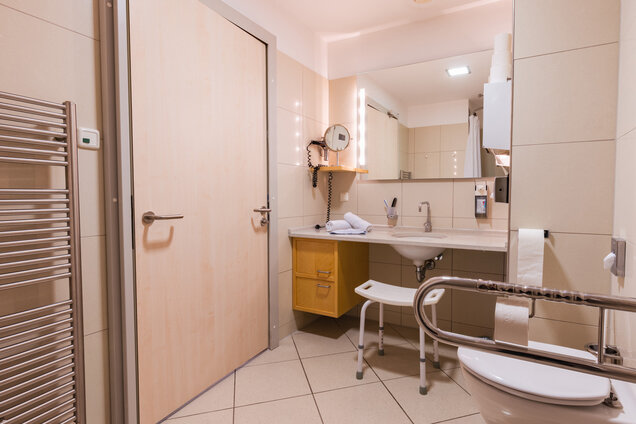 Das Bild zeigt ein modernes Badezimmer mit einem Waschbecken, einem Spiegel und einem Toilettenbereich. Es gibt einen Handtuchwärmer, eine Holzkommode mit zwei Schubladen und eine Stuhleinrichtung für zusätzlichen Komfort. Die Wände sind hell gefliest und es gibt einen großen Spiegel über dem Waschbecken.