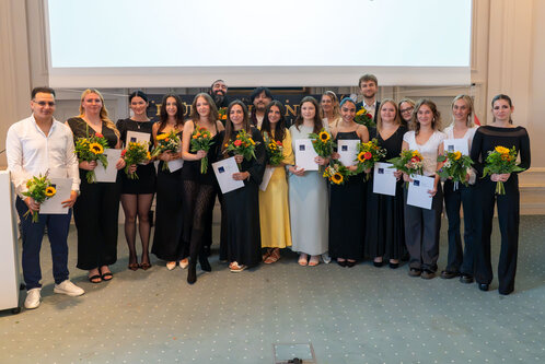 Eine Gruppe von 15 Personen steht auf einer Bühne, alle mit Blumensträußen in den Händen. Sie tragen festliche Kleidung und lächeln. Im Hintergrund ist eine Leinwand zu sehen. Die Aufstellung vermittelt eine feierliche Stimmung, möglicherweise eine Abschlussfeier oder Auszeichnung.