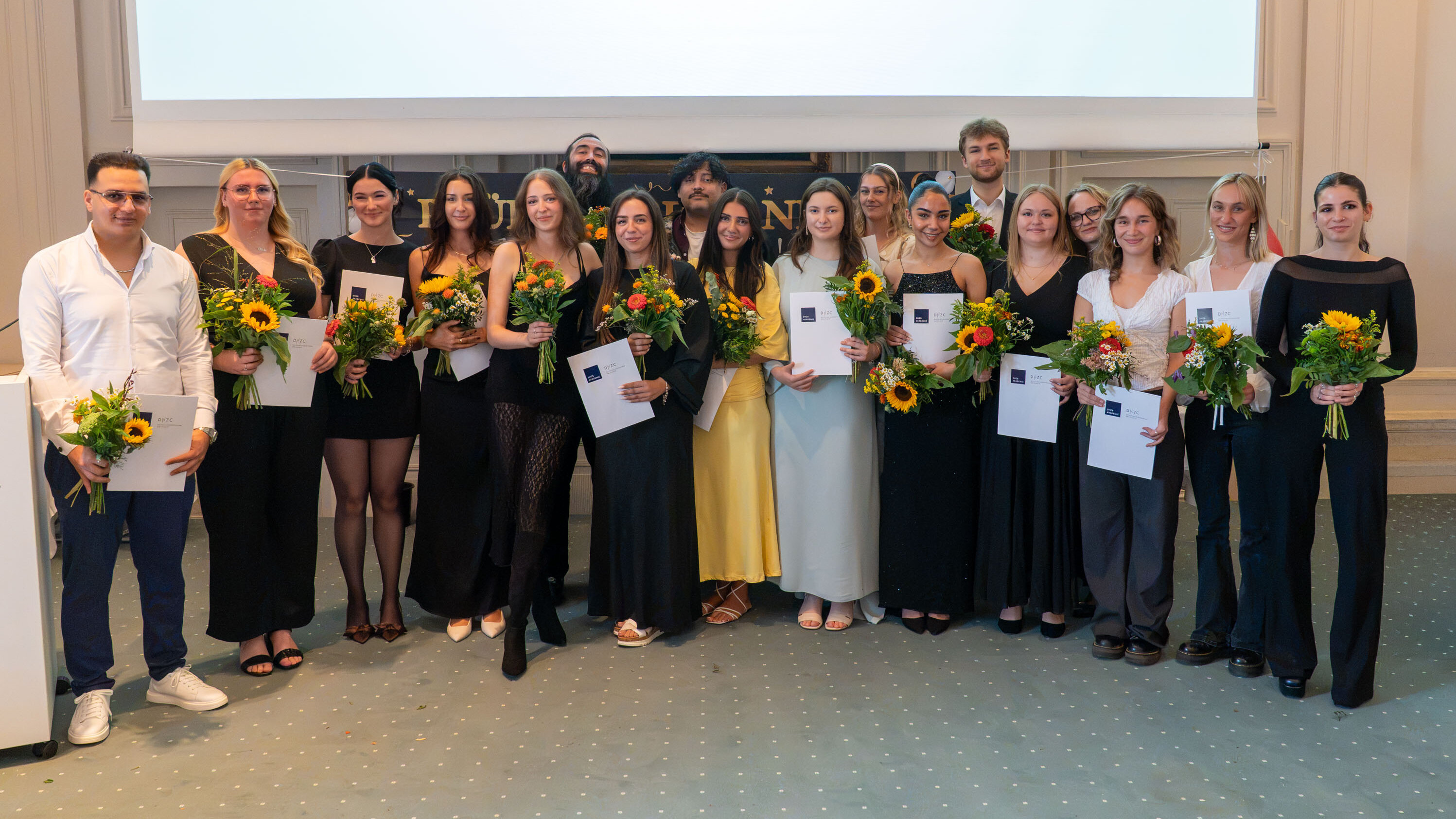 Eine Gruppe von 15 Personen steht auf einer Bühne, alle mit Blumensträußen in den Händen. Sie tragen festliche Kleidung und lächeln. Im Hintergrund ist eine Leinwand zu sehen. Die Aufstellung vermittelt eine feierliche Stimmung, möglicherweise eine Abschlussfeier oder Auszeichnung.