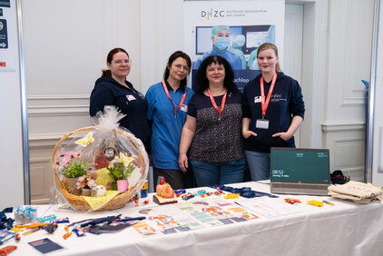 Auf dem Bild sind vier Frauen zu sehen, die an einem Informationsstand stehen. Sie tragen verschiedene T-Shirts und Lanyards. Vor ihnen befindet sich ein Tisch mit Werbematerialien, Süßigkeiten und einem Körbchen mit Geschenken. Im Hintergrund ist ein Banner mit dem Logo und dem Namen der Organisation zu erkennen.