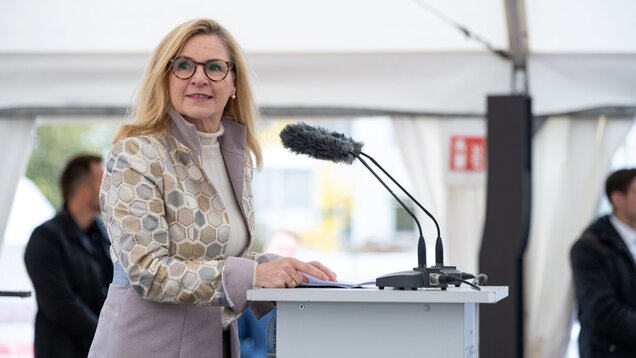 Eine Frau mit blonden Haaren und Brille steht an einem Rednerpult, das ein Mikrofon hat. Sie trägt einen hellen, strukturierten Mantel und schaut aufmerksam. Im Hintergrund sind verschwommene Umrisse von Personen und einem Zelt zu erkennen.