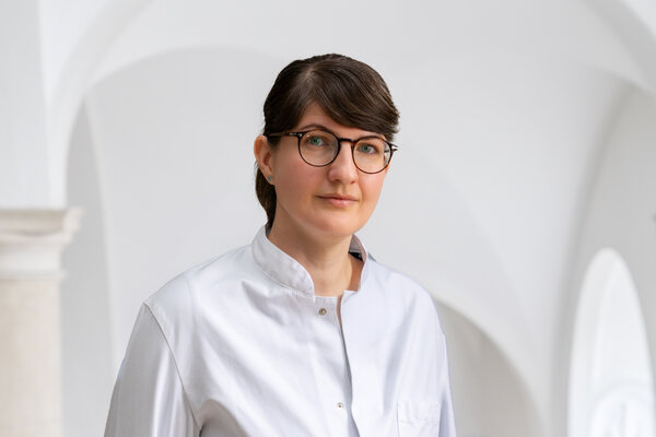Eine Frau mit kurzen, braunen Haaren und einer Brille trägt eine weiße Bluse. Sie steht in einem hellen Raum mit einer minimalistischen Hintergrundgestaltung. Ihr Ausdruck ist neutral und professionell.
