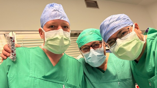 Drei Ärzte in OP-Kleidung stehen eng zusammen. Sie tragen Gesichtsmasken und chirurgische Hauben in verschiedenen Farben. Alle lächeln in die Kamera. Im Hintergrund sind neutrale Krankenhauswände zu sehen, was die medizinische Umgebung unterstreicht.