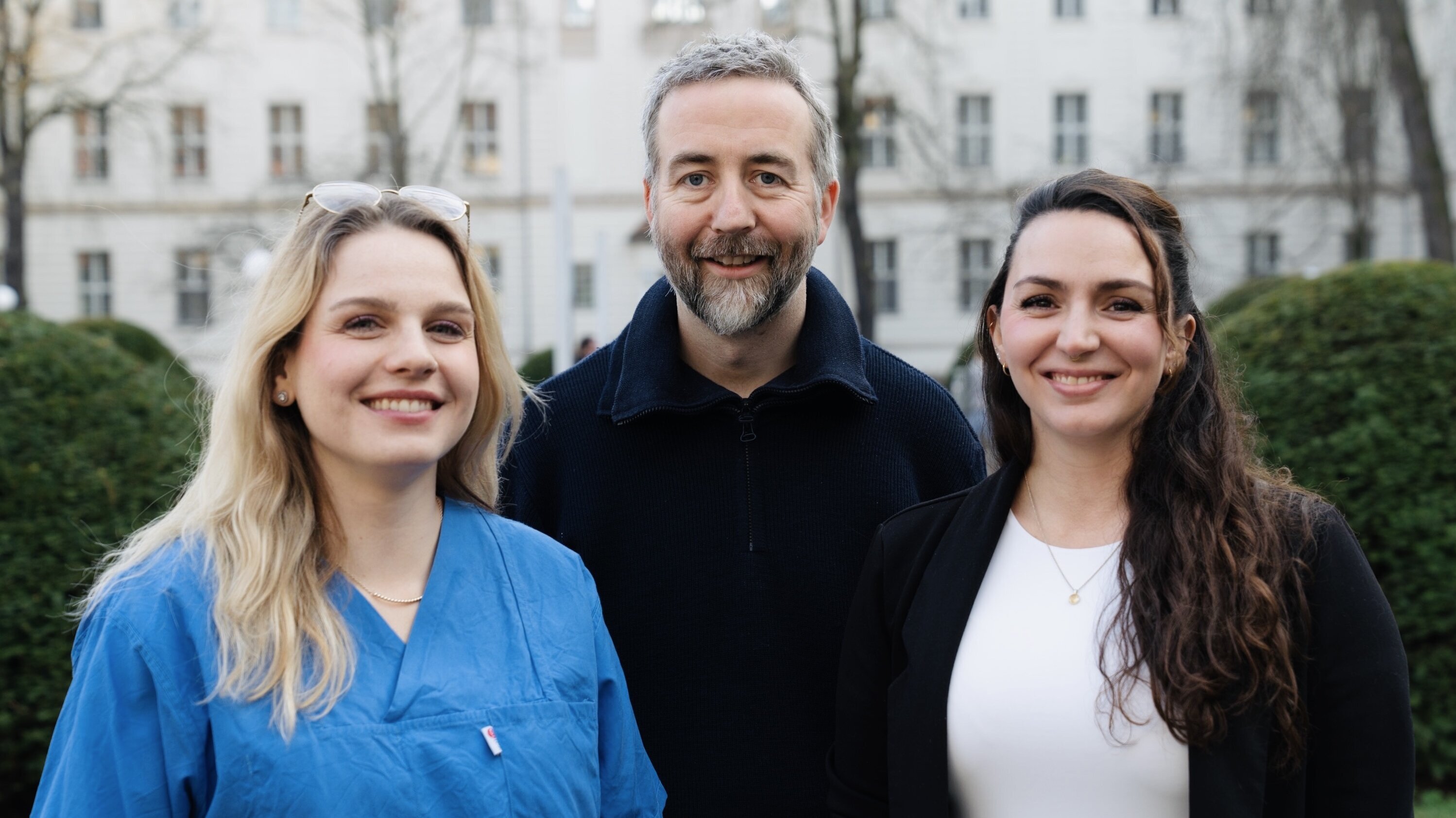 Drei Personen stehen im Freien, mit Bäumen und Gebäuden im Hintergrund. Die Frau links trägt ein blaues Oberteil, der Mann in der Mitte hat graues Haar und einen Bart, die Frau rechts trägt ein weißes Top. Alle lächeln freundlich.