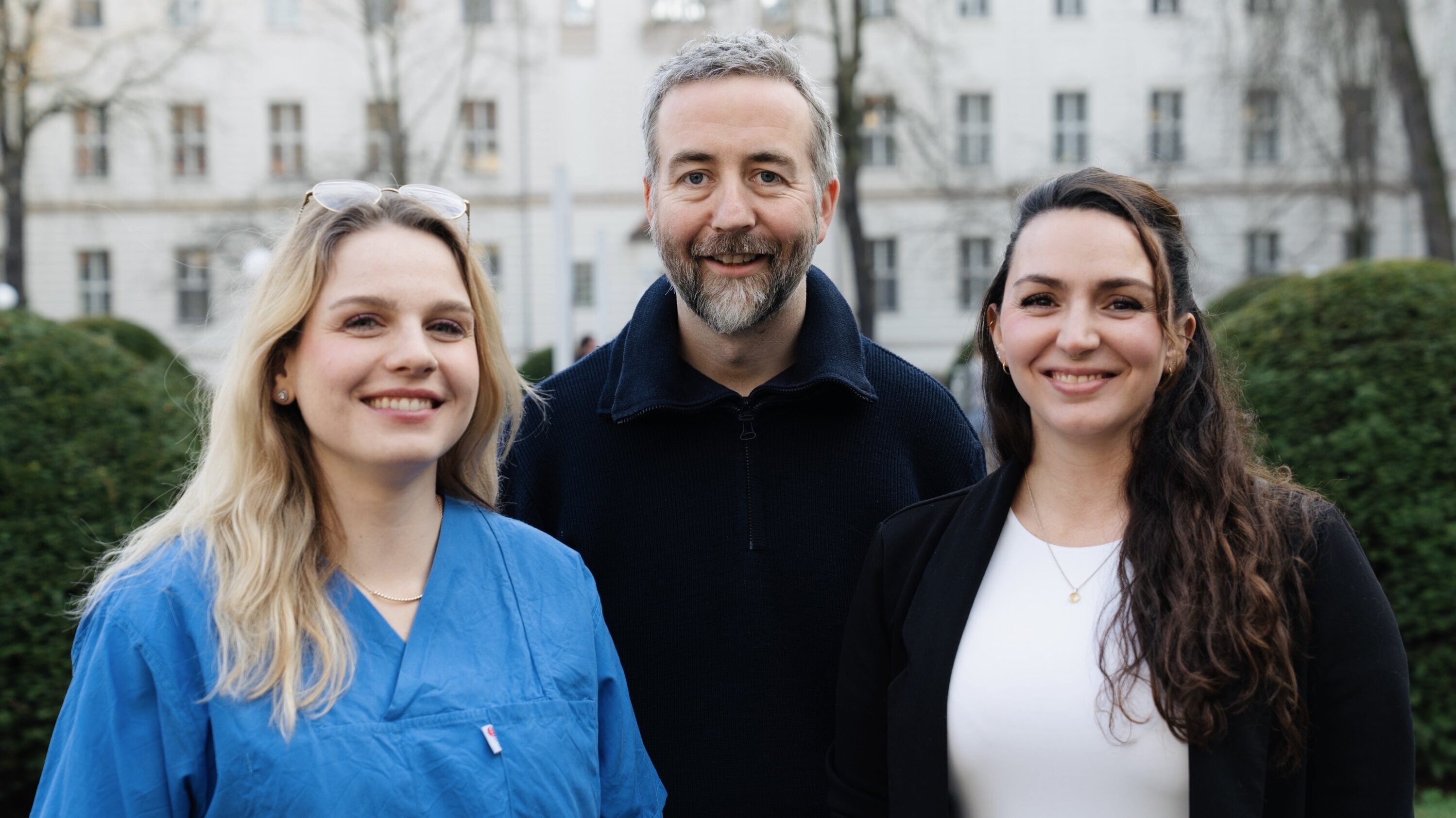Drei Personen stehen im Freien, mit Bäumen und Gebäuden im Hintergrund. Die Frau links trägt ein blaues Oberteil, der Mann in der Mitte hat graues Haar und einen Bart, die Frau rechts trägt ein weißes Top. Alle lächeln freundlich.