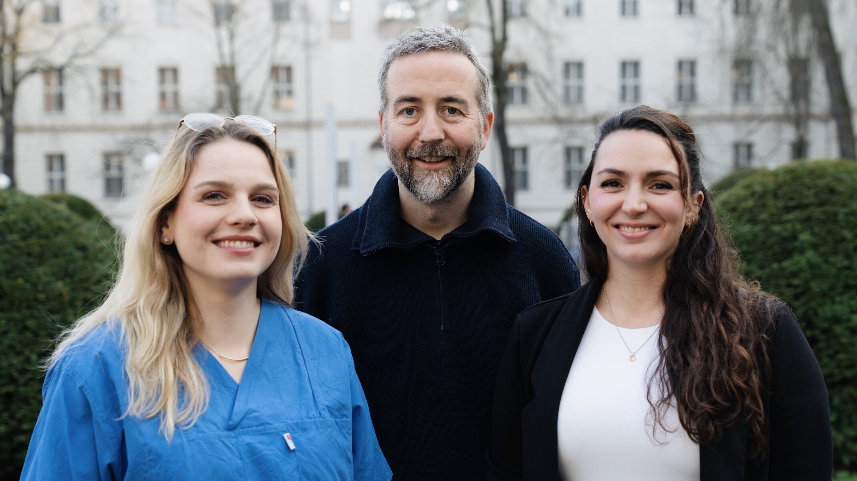 Drei Personen stehen im Freien, mit Bäumen und Gebäuden im Hintergrund. Die Frau links trägt ein blaues Oberteil, der Mann in der Mitte hat graues Haar und einen Bart, die Frau rechts trägt ein weißes Top. Alle lächeln freundlich.