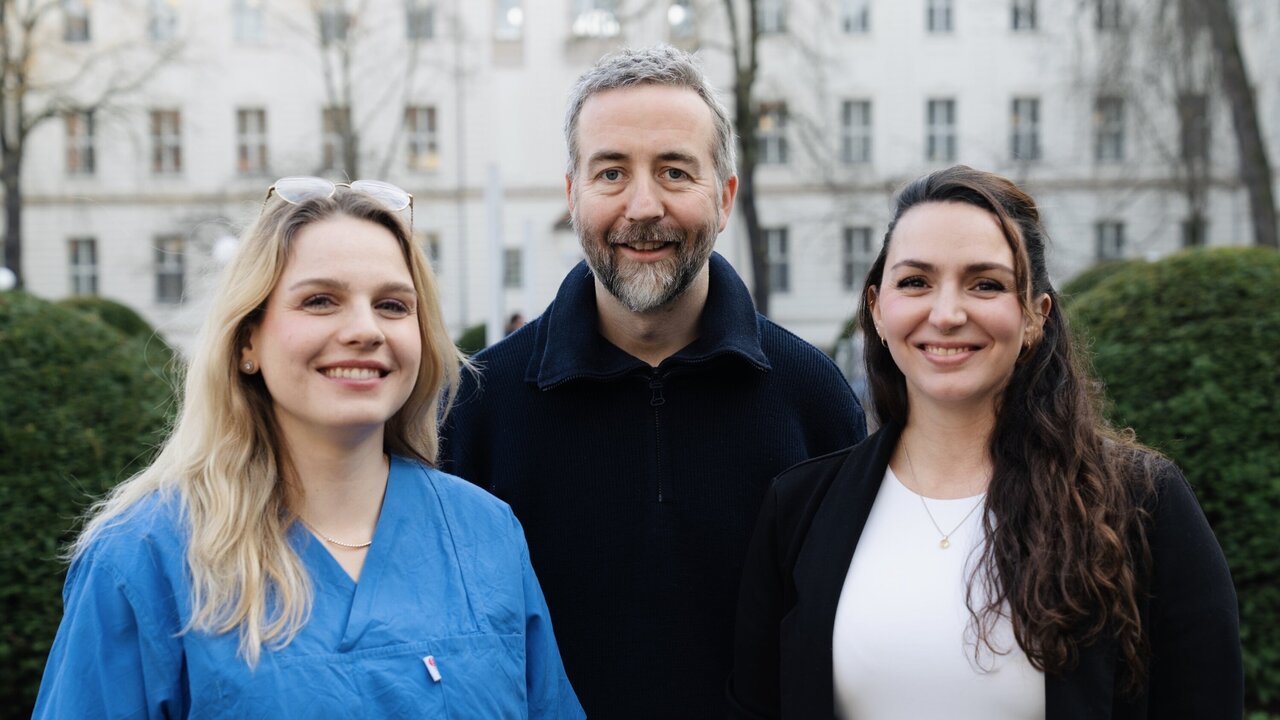 Drei Personen stehen im Freien, mit Bäumen und Gebäuden im Hintergrund. Die Frau links trägt ein blaues Oberteil, der Mann in der Mitte hat graues Haar und einen Bart, die Frau rechts trägt ein weißes Top. Alle lächeln freundlich.