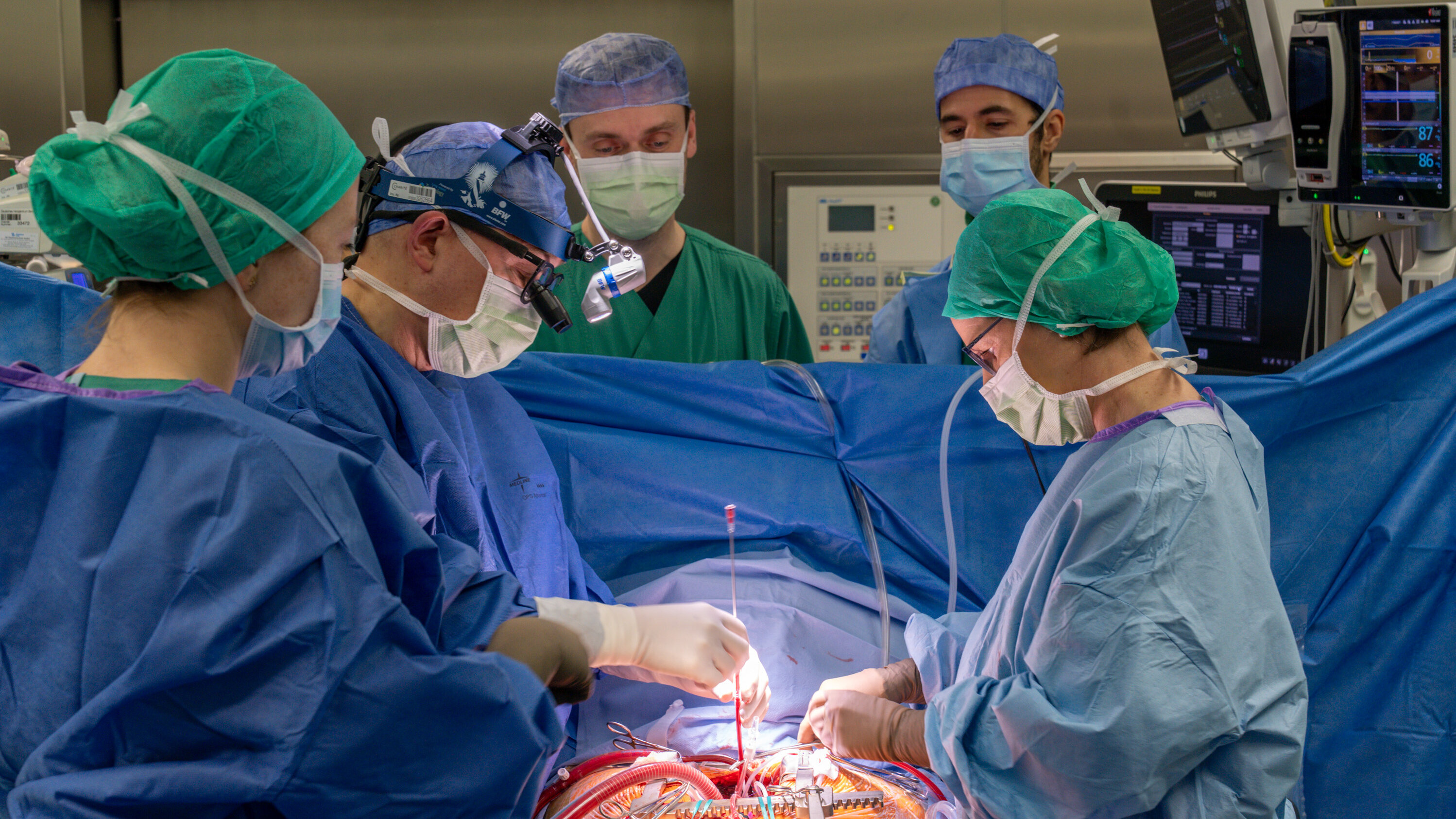 In einem Operationssaal sind mehrere Chirurgen und medizinisches Personal in blauer und grüner Sterilkleidung zu sehen. Sie führen eine komplexe Operation durch, während sie sich konzentriert um den Patienten kümmern, der auf dem Operationstisch liegt.
