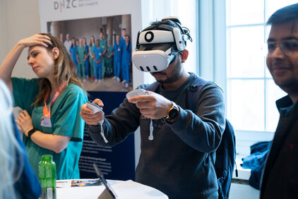 Eine Person trägt eine VR-Brille und interagiert mit einer virtuellen Umgebung, während sie zwei Controller in den Händen hält. Im Hintergrund sind Personen in medizinischer Kleidung und ein Informationsstand zu sehen. Der Fokus liegt auf der virtuellen Erfahrung des Nutzers.