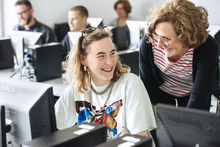 Eine lächelnde junge Frau sitzt an einem Computer, während eine ältere Frau hinter ihr steht und freundlich mit ihr spricht. Beide wirken fröhlich und gleichzeitig konzentriert. Im Hintergrund sind weitere Personen an ihren Computern zu sehen.