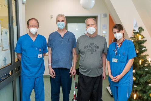 Vier Personen stehen in einem Krankenhausflur, alle tragen Atemschutzmasken. Zwei der Personen sind medizinisches Personal in blauer Kleidung, während zwei weitere Patienten in Freizeitkleidung sind. Im Hintergrund ist ein Weihnachtsbaum mit Dekorationen zu sehen.