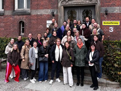 Eine Gruppe von etwa 40 Personen steht vor einem historischen Gebäude mit einer kunstvollen Eingangstür. Die Teilnehmer lächeln und posieren zusammen, um an einem gemeinsamen Ereignis oder Treffen teilzunehmen. Das Gebäude ist von Pflanzen umgeben und hat große Fenster.