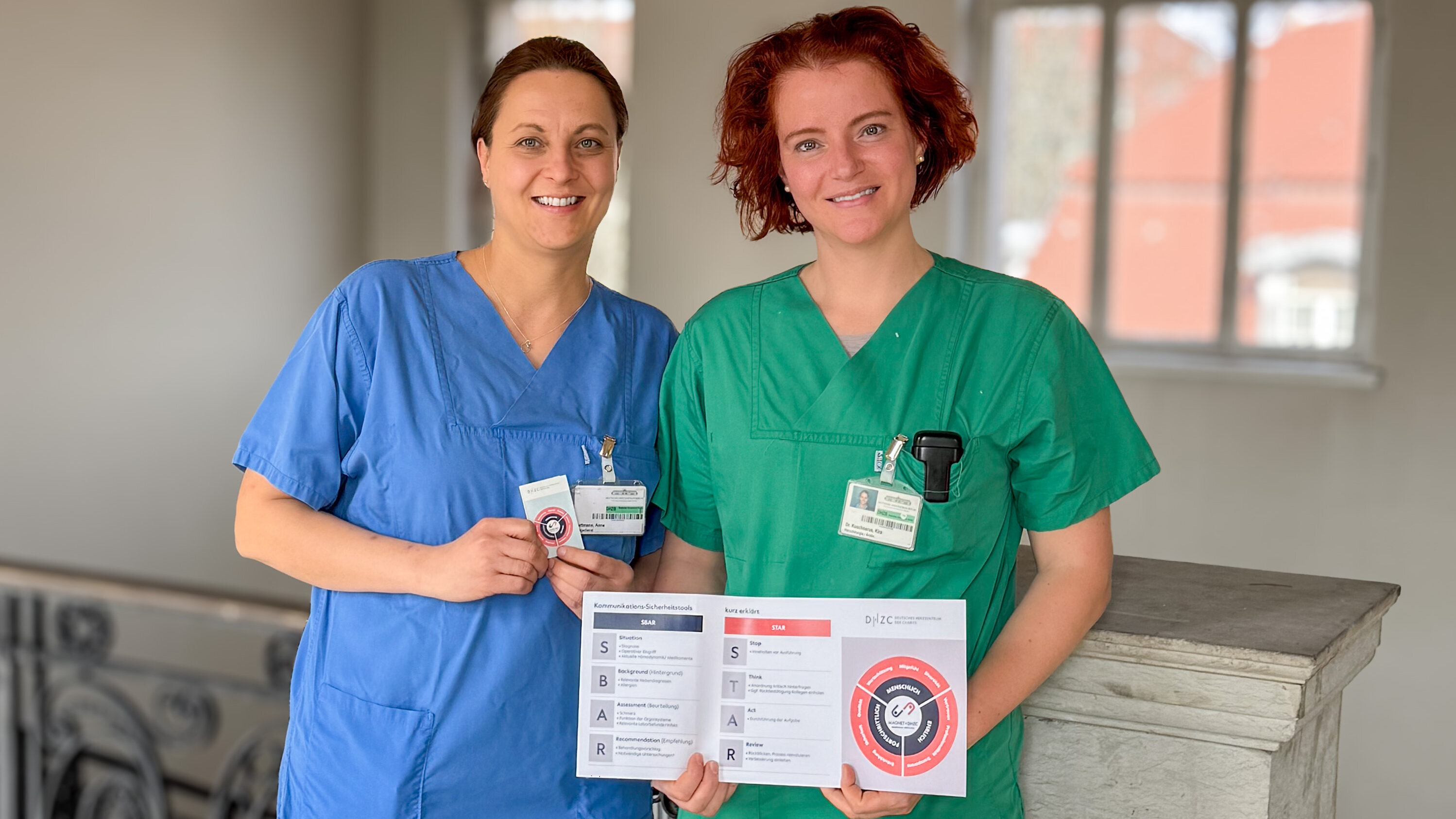 Zwei medizinische Fachkräfte stehen in einem hellen, offenen Raum. Die eine trägt ein blaues, die andere ein grünes Pflegeoutfit. Beide halten ein Informationsblatt, das verschiedene Kommunikationsmethoden zeigt. Sie lächeln und strahlen Professionalität und freundliche Zusammenarbeit aus.