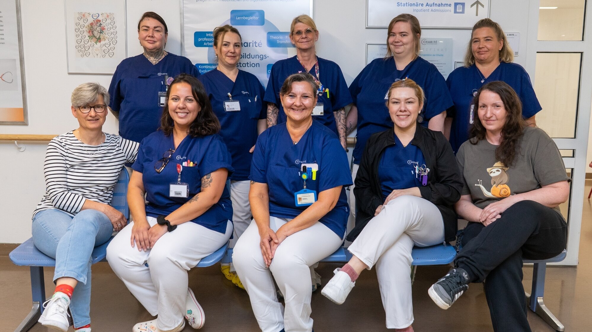 Eine Gruppe von elf Personen in blauen Krankenpflegeuniformen posiert lächelnd in einem Raum. Sie sitzen auf Bänken und stehen hintereinander. Im Hintergrund hängt ein Willkommen-Schild. Die Atmosphäre wirkt freundlich und einladend.