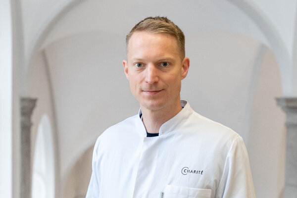 Ein Arzt in einem weißen Kittel steht vor einem neutralen Hintergrund. Er hat kurzes, helles Haar und schaut direkt in die Kamera. Auf seinem Kittel ist das Logo der Charité zu sehen.