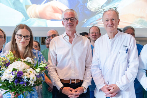 Eine Gruppe von medizinischem Personal steht zusammen, darunter eine Frau mit Blumenstrauß, ein Mann in Hemd und Kragen, und ein Arzt in weißen Kittel. Im Hintergrund ist eine Präsentation mit einer medizinischen Szene sichtbar. Alle wirken glücklich und stolz.