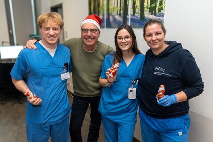 Vier Personen stehen zusammen, lächeln und halten Schokoladenfiguren in den Händen. Zwei tragen blaue Mitarbeiteruniformen, eine Person hat einen Weihnachtsmann-Hut auf, und die vierte Person trägt ein schwarzes Sweatshirt mit einem Logo. Im Hintergrund ist eine Wand mit einem Gemälde von Bäumen.