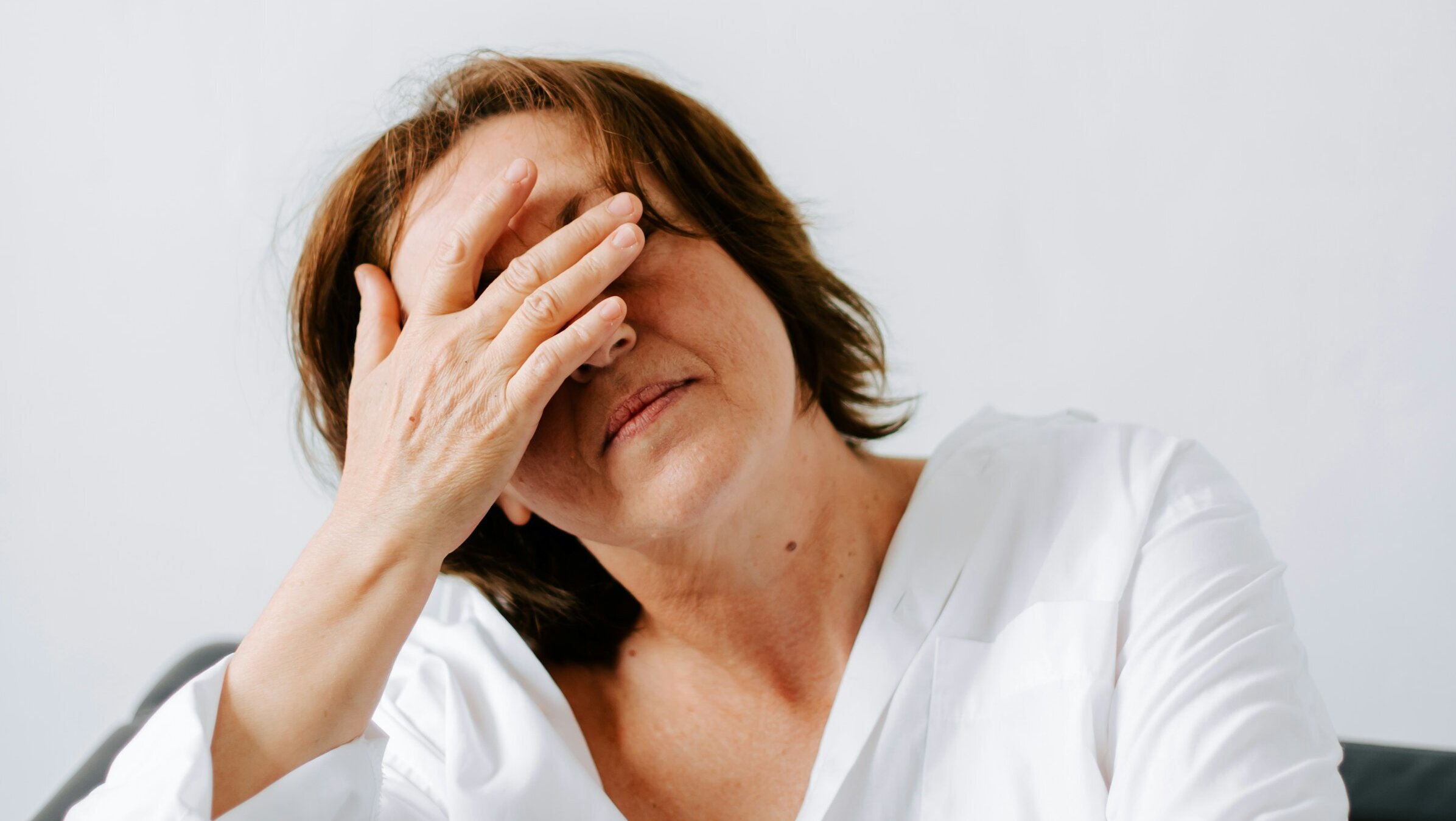 Eine Frau sitzt auf einer Couch und hält eine Hand an ihr Gesicht. Sie trägt eine weiße Bluse und scheint nachdenklich oder gestresst zu sein. Der Hintergrund ist hell und schlicht, was ihre Gemütsverfassung verstärkt.