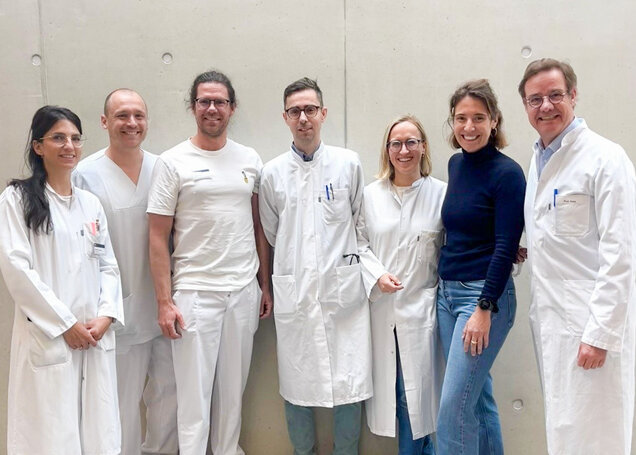 Eine Gruppe von sieben Personen in weißen Kitteln steht lächelnd vor einer hellen, glatten Wand. Sie sind medizinisches Fachpersonal, darunter Ärzte und Pflegekräfte, und bilden eine Einheit, die Teamarbeit und Professionalität ausstrahlt.