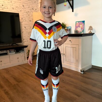 Ein Mädchen steht in einem Wohnzimmer in einem Fußballtrikot der deutschen Nationalmannschaft. Sie trägt ein Trikot mit den Farben Schwarz, Rot und Gold, eine schwarze Shorts und Socken mit DFB-Aufdruck. Sie lächelt und posiert mit einer Hand an der Hüfte.