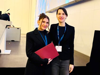 Zwei Frauen stehen auf einer Bühne, die eine hält eine rote Mappe. Beide tragen schwarze Blazer und lächeln. Im Hintergrund sind unscharfe Elemente einer Veranstaltung zu erkennen.