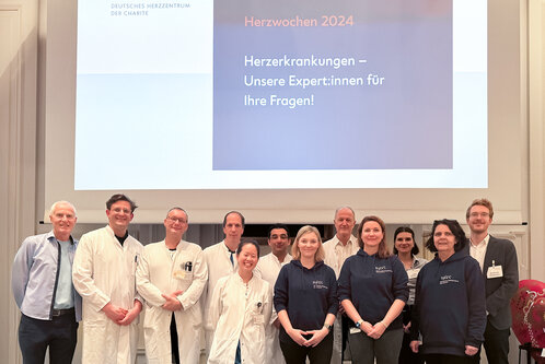 Eine Gruppe von Fachleuten in medizinischer Kleidung steht vor einem Bildschirm mit Informationen zu Herzwochen 2024. Der Text auf dem Bildschirm thematisiert Herzerkrankungen und betont die Expertise der Präsentierenden für Fragen zu diesem Thema.