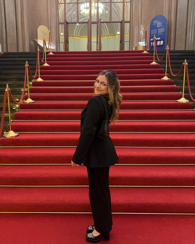 Eine lächelnde Frau trägt einen eleganten schwarzen Anzug und posiert auf einer roten Teppichtreppe. Die Treppe ist von goldenen Stangen und roten Seilen gesäumt, im Hintergrund sind große Glastüren sichtbar.