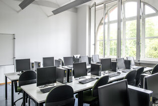 Ein Raum mit mehreren Computern auf Tischen. Jedes Arbeitsplätzchen besteht aus einem Bildschirm, einer Tastatur und einem Computergehäuse. Große Fenster ermöglichen viel Tageslicht und bieten einen Blick nach draußen. Der Raum wirkt modern und einladend.