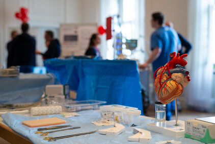 Das Bild zeigt einen Tisch mit medizinischen Instrumenten und Ausstattungen, auf dem ein Modell eines menschlichen Herzens prominent steht. Im Hintergrund sind Personen zu sehen, die sich unterhalten. Die Atmosphäre scheint eine medizinische Veranstaltung oder Präsentation zu sein.