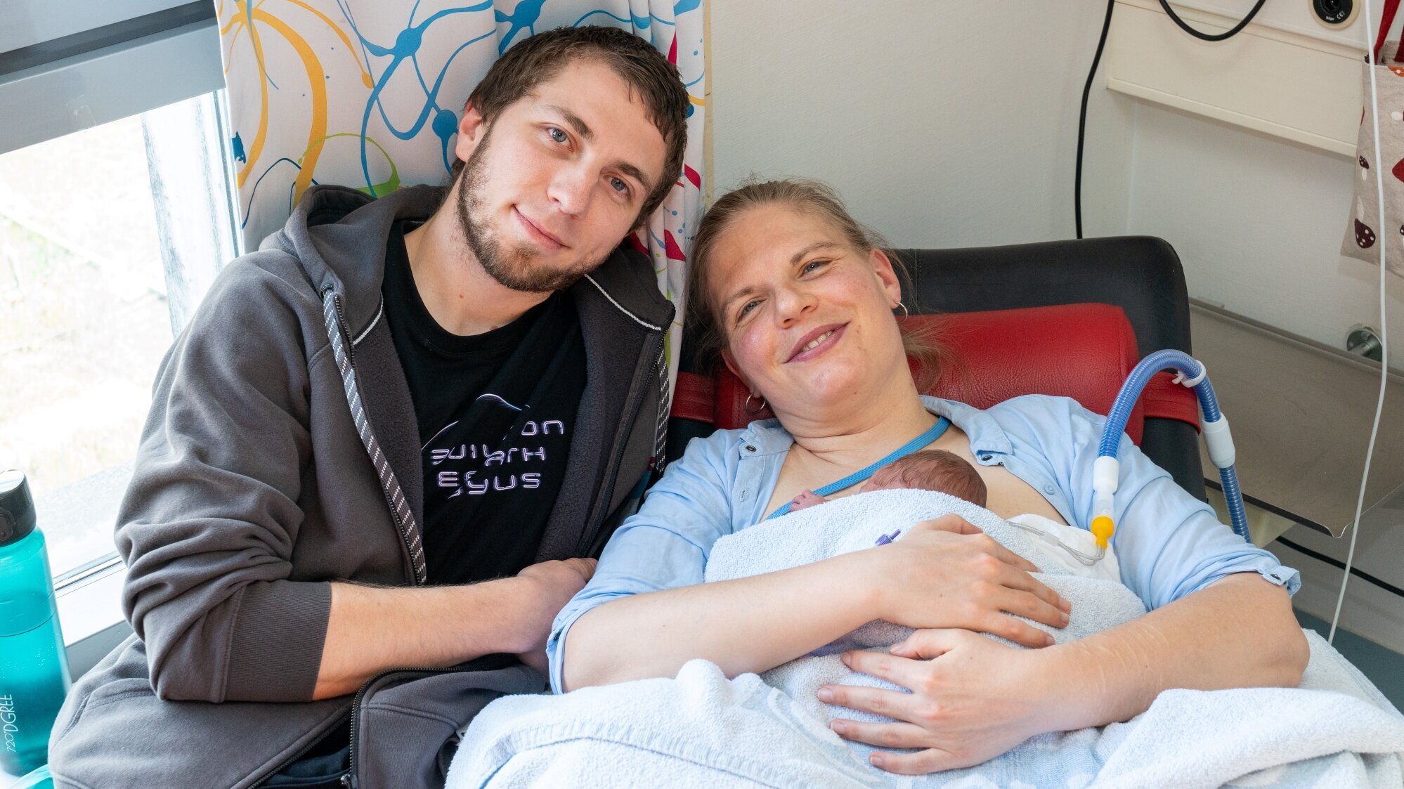Eine Mutter liegt auf einem Krankenhausbett und hält ihr Neugeborenes in den Armen. Neben ihr sitzt der Vater, der sie anlächelt. Beide strahlen Freude und Zuneigung aus, während sie diesen besonderen Moment gemeinsam erleben.