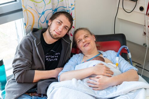 Eine Mutter liegt auf einem Krankenhausbett und hält ihr Neugeborenes in den Armen. Neben ihr sitzt der Vater, der sie anlächelt. Beide strahlen Freude und Zuneigung aus, während sie diesen besonderen Moment gemeinsam erleben.