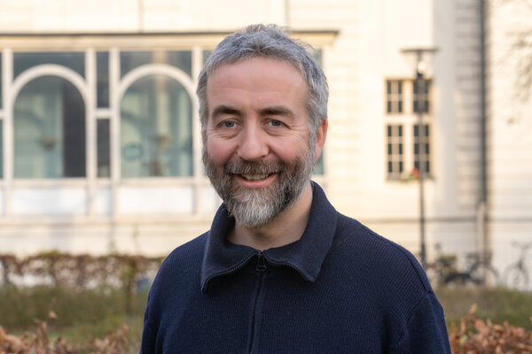 Ein lächelnder Mann mit grauen Haaren und einem Bart steht in einem Park. Er trägt einen blauen Pullover mit Reißverschluss. Im Hintergrund sind Fenster und Pflanzen zu sehen, die zur umgebenden Architektur gehören.
