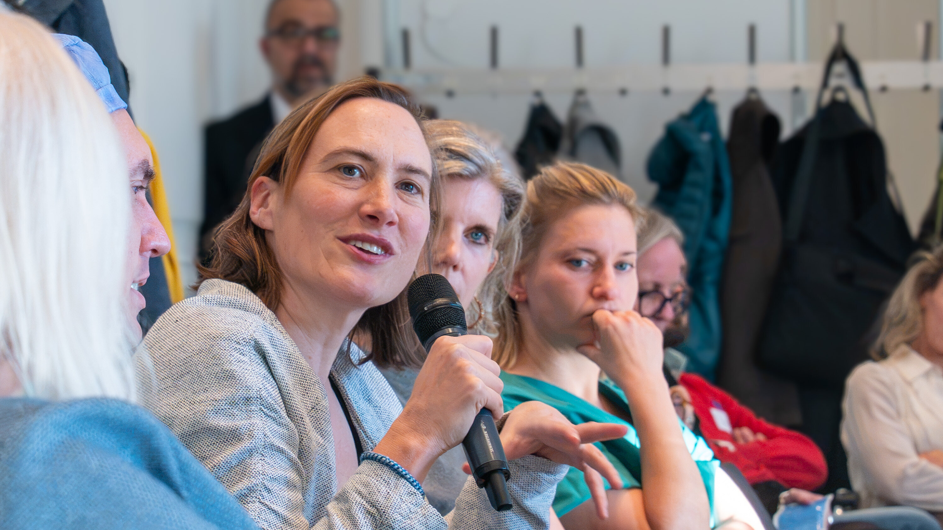 Eine Frau spricht in ein Mikrofon, während sie in einer Diskussionsrunde sitzt. Ihre Gestik deutet auf Engagement hin. Im Hintergrund sind weitere Teilnehmer zu sehen, die aufmerksam zuhören.