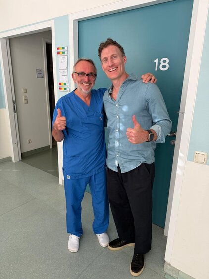 Zwei Männer stehen in einem Krankenhaussaal vor einer Tür mit der Nummer 18. Der Mann links trägt einen blauen Kittel und hat eine Brille, während der Mann rechts in einer blauen Hemd und schwarzen Hose gekleidet ist. Beide zeigen ein positives Daumen-hoch-Zeichen.