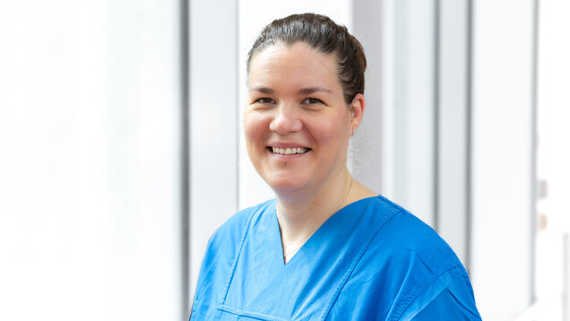 Eine lächelnde Frau in blauer medizinischer Kleidung steht vor großen Fenstern. Sie hat kurze, braune Haare und wirkt freundlich und professionell.