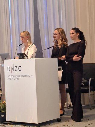 Drei Frauen stehen an einem Rednerpult im Deutschen Herz-Zentrum der Charité. Eine Frau spricht ins Mikrofon, während die anderen beiden aufmerksam zuhören. Sie sind formell gekleidet und auf einer Veranstaltung, die vermutlich eine Präsentation oder ein Seminar beinhaltet.