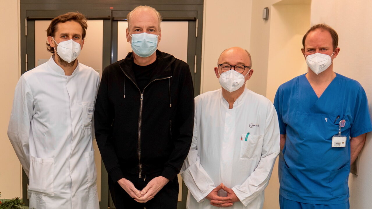 Vier Personen stehen nebeneinander in einem Krankenhausflur. Sie tragen Masken; zwei in weißen Ärztekitteln, einer in blauer Arbeitskleidung und eine Person in schwarzem Oberteil. Im Hintergrund ist eine Tür und eine helle Lampe zu sehen.