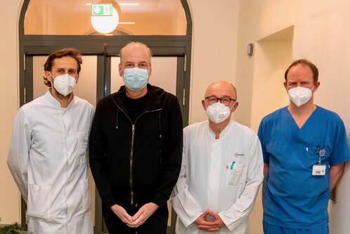 Vier Personen stehen nebeneinander in einem Krankenhausflur. Sie tragen Masken; zwei in weißen Ärztekitteln, einer in blauer Arbeitskleidung und eine Person in schwarzem Oberteil. Im Hintergrund ist eine Tür und eine helle Lampe zu sehen.