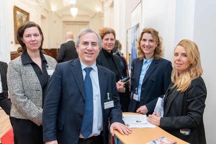 Eine Gruppe von fünf Personen steht in einem Hellen Flur. Alle sind bekleidet und lächeln. Im Vordergrund steht ein Mann in einem Anzug mit einer Krawatte. Auf dem Tisch vor ihnen liegen Dokumente.