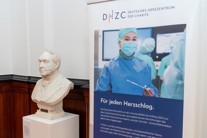 Das Bild zeigt eine Informationswand des Deutschen Herzzentrums der Charité (DHZC) mit einer abgebildeten Ärztin in Schutzkleidung. Neben der Wand steht eine Büste eines Mannes. Der Banner betont die Bedeutung des DHZC für die Herzgesundheit.