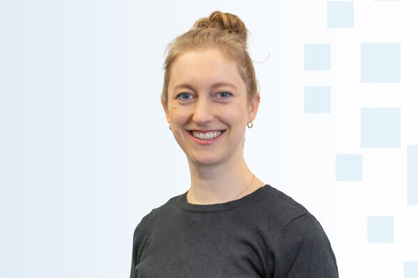 Eine lächelnde Frau mit blonden Haaren, die zu einem Dutt gebunden sind, trägt einen dunkelgrauen Pullover. Der Hintergrund ist hell und besteht aus hellen geometrischen Formen.