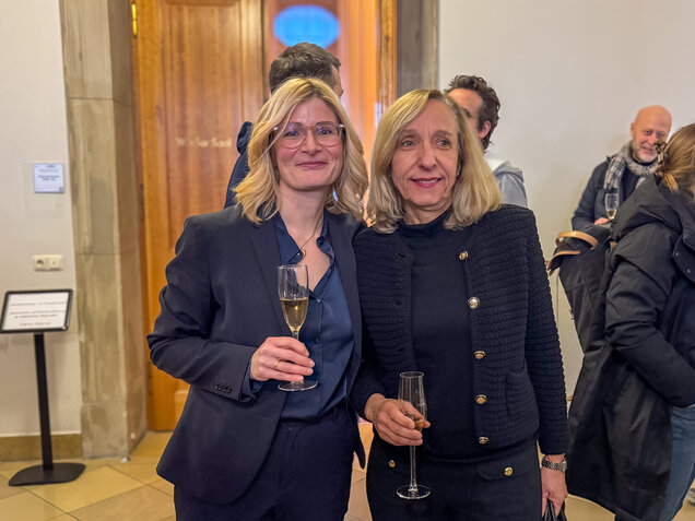 Zwei Frauen stehen lächelnd nebeneinander, jede hält ein Glas Sekt. Die Frau links trägt einen dunklen Anzug, die Frau rechts eine schwarze Jacke. Im Hintergrund sind weitere Personen und ein Holztür zu sehen.