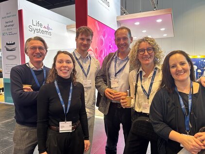 Sechs Personen stehen zusammen auf einer Messe, freundlich lächelnd. Sie tragen Namensschilder und halten Tassen in den Händen. Im Hintergrund sind Stände mit Werbeplakaten zu sehen. Die Gruppe strahlt eine positive Atmosphäre aus und steht in einem modernen Messeumfeld.