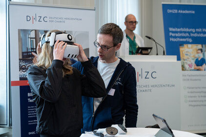 Eine Person trägt eine Virtual-Reality-Brille und wird von einer zweiten Person angeleitet. Im Hintergrund sind Informationsstände des Deutschen Herzzentrums zu sehen, die über Bildungsangebote informieren.